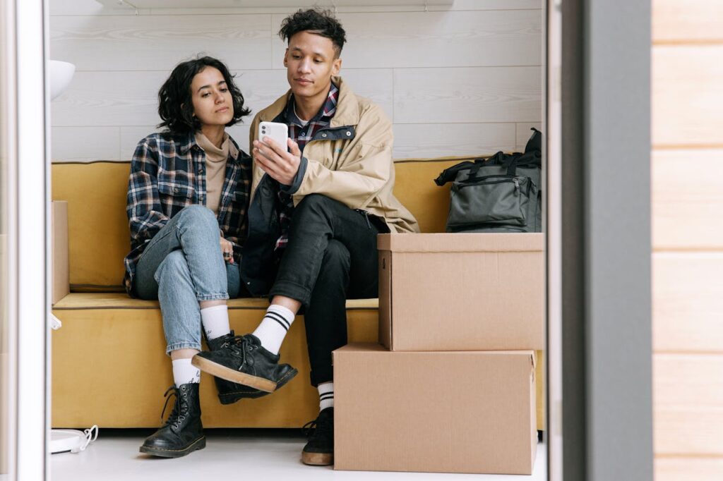 Young couple sitting on sofa among moving boxes, using smartphones in their new home.